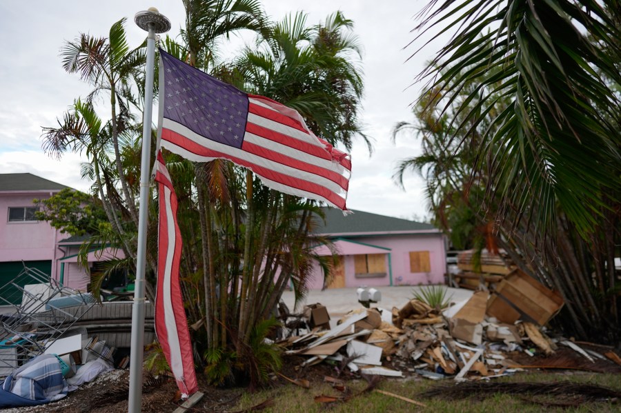FILE - A tattered American flag flaps outside a home as furniture and household items damaged by Hurricane Helene flooding sit piled along the street awaiting pickup, ahead of the arrival of Hurricane Milton, in Holmes Beach on Anna Maria Island, Fla., Tuesday, Oct. 8, 2024. (AP Photo/Rebecca Blackwell, File)