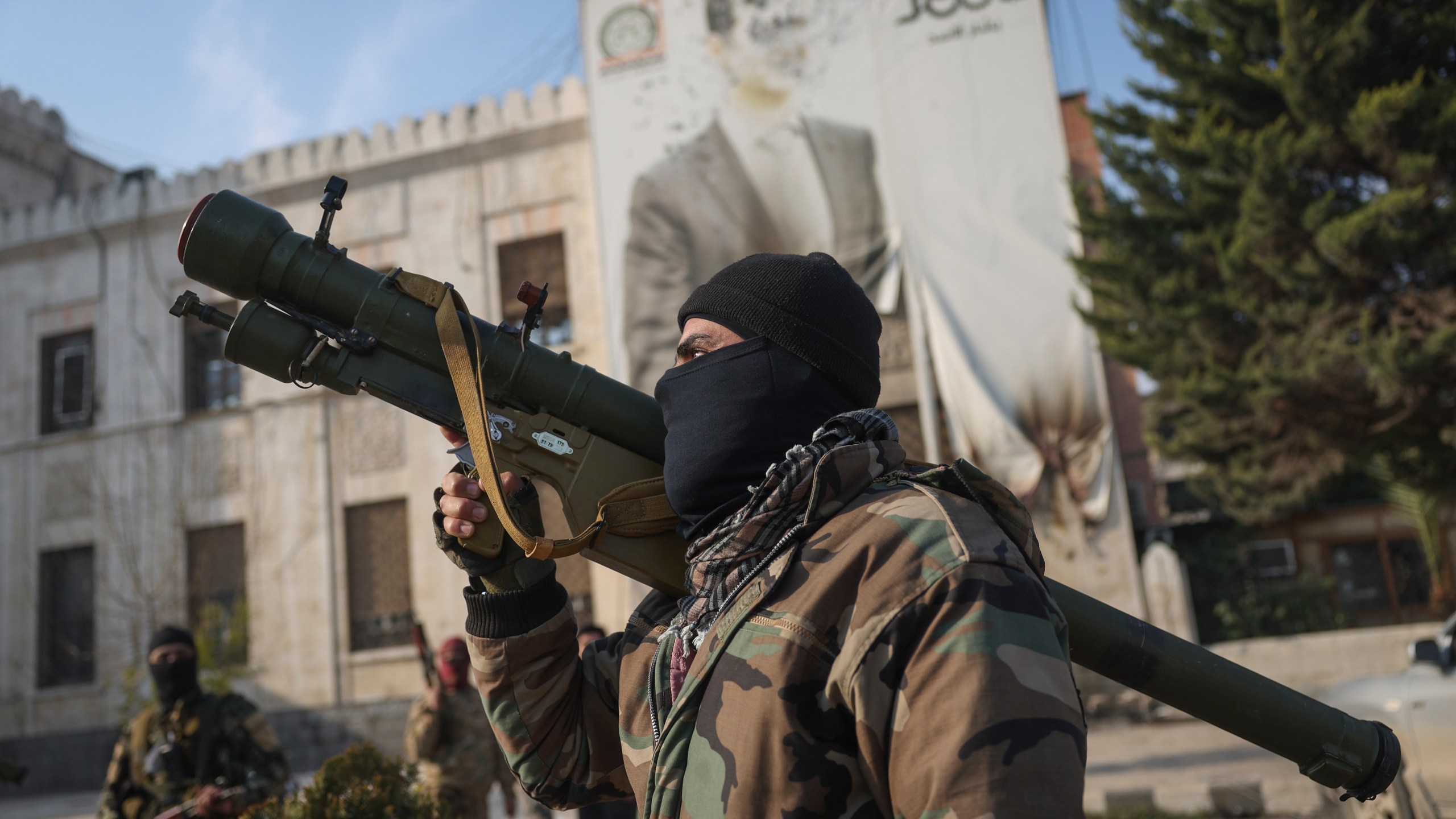 A Syrian opposition fighter holds a rocket launcher in front of the provincial government office, where an image of Syrian President Bashar Assad is riddled with bullets on the facade, in the aftermath of the opposition's takeover of Hama, Syria, Friday, Dec. 6, 2024. (AP Photo/Ghaith Alsayed)